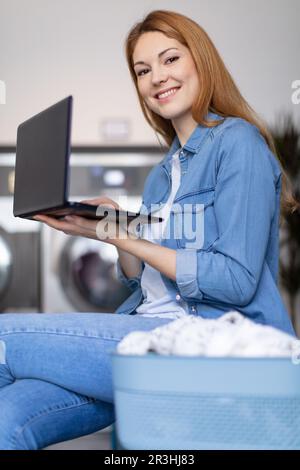 Nette Frau, die die Waschmaschine überprüft, während sie mit dem Laptop arbeitet Stockfoto