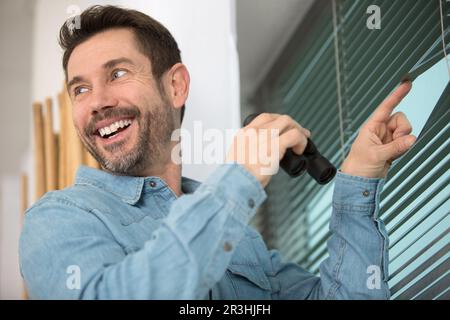 Ein glücklicher Mann, der mit einem Fernglas durch die Jalousien guckt Stockfoto