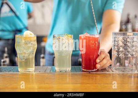 Drei sommerliche Cocktails in Steingläsern am Rand einer Bar, wobei der Barkeeper nach einem greift Stockfoto