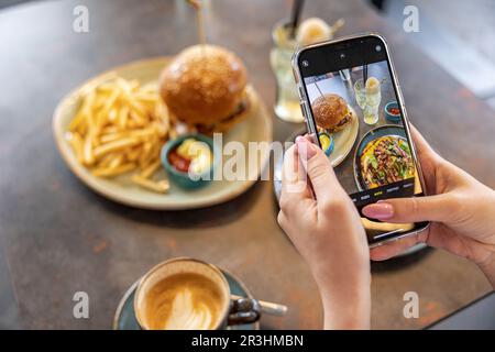 Draufsicht einer jungen Frau, die mit dem Smartphone im Heimstudio ein ästhetisches Foto von Lebensmitteln macht. Foto von Burger und Pommes frites Stockfoto