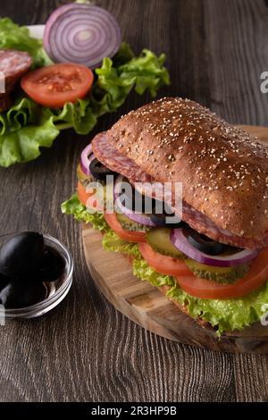 Foto-Sandwich mit Wurst, Oliven, Tomaten, Salatblättern und Gurken auf einem Holzbrett, vertikales Foto Stockfoto