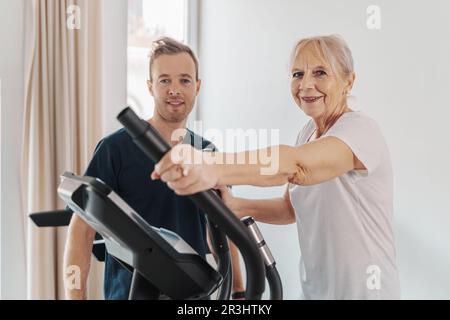 Seniorin mit Personal Trainer im Fitnessstudio: Eine lächelnde Seniorin, die unter Anleitung ihres Personal Trainers auf einer Stepper trainiert. Beide Augen Stockfoto