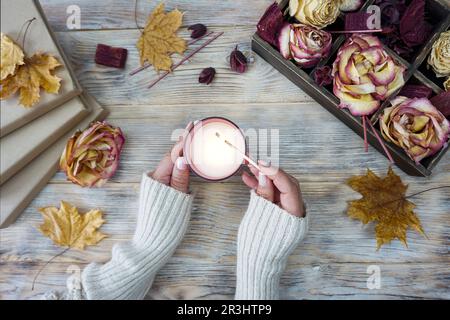 Eine Frau zündet eine aromatische Kerze an. Auf dem Tisch liegen getrocknete Rosen und gelbe Ahornblätter. Stockfoto