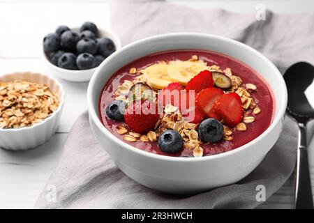 Köstliche Smoothie-Schüssel mit frischen Beeren, Banane und Müsli auf weißem Holztisch, Nahaufnahme Stockfoto