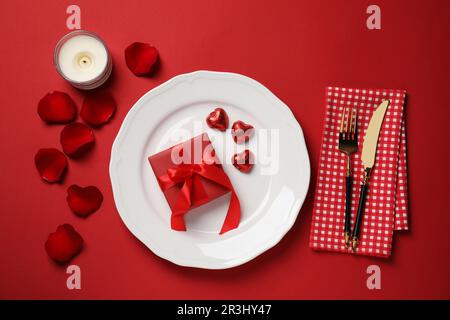 Wunderschöne Tischgestaltung mit Geschenkbox und Rosenblättern zum Abendessen auf einem roten Tisch für romantische, flache Bettwäsche Stockfoto