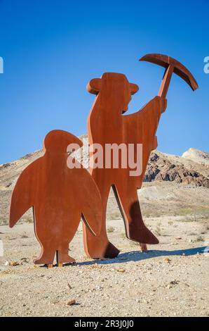 Eine rostige Stahlausschnittfigur eines Hutmachers, der eine Spitzhacke erhebt, steht neben einer Pinguinausschnittfigur auf hellem Wüstenboden in der Nähe von Rhyolite, Nevada. Stockfoto