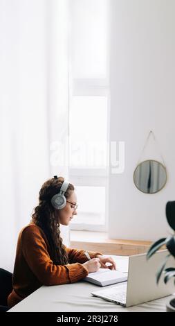 Junger Student, der ein virtuelles Seminar besucht, während er zu Hause studiert. Frau trägt Kopfhörer, schreibt auf dem Laptop und macht sich während des E-Learning Notizen Stockfoto