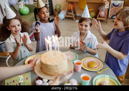 Blick aus einem hohen Winkel auf eine vielfältige Gruppe glücklicher Kinder, die klatschen, während sie auf den Kuchen mit Kerzen während der Geburtstagsparty warten Stockfoto