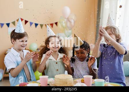 Vielfältige Gruppe von aufgeregten kleinen Kindern, die Konfetti werfen, während sie eine Geburtstagsparty für Kinder genießen und Spaß haben Stockfoto