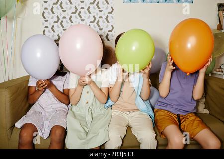 Eine vielfältige Gruppe von Kindern hält Ballons und versteckt Gesichter während der Party für Kinder, die mit Blitz aufgenommen wurden Stockfoto