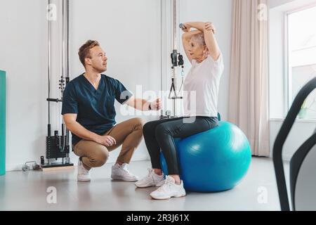Seniorin sitzt auf einem Pilates-Ball im Fitnessstudio, während ein Personal Trainer Trainingsprogramm durchführt. Sowohl lächelnd als auch engagiert beim Workout. Stockfoto