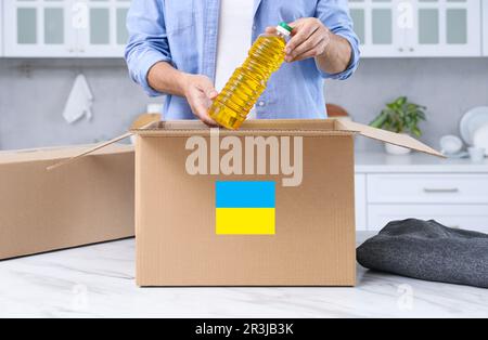 Humanitäre Hilfe für die Ukraine. Ein Mann, der eine Flasche Öl in eine Spendenbox am weißen Tisch im Haus legt, Nahaufnahme Stockfoto