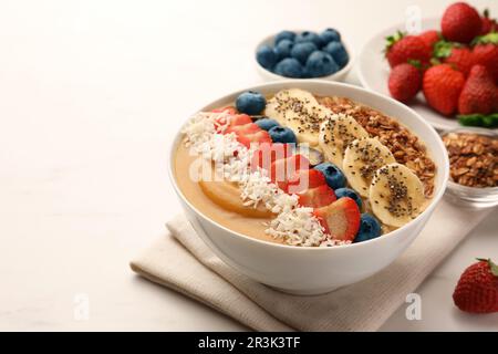 Köstliche Smoothie-Schüssel mit frischen Beeren, Banane, Kokosflocken und Müsli auf dem weißen Tisch. Platz für Text Stockfoto