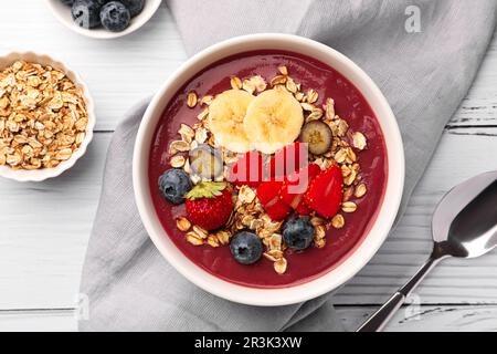 Köstliche Smoothie-Schüssel mit frischen Beeren, Banane und Müsli auf weißem Holztisch, flach liegend Stockfoto