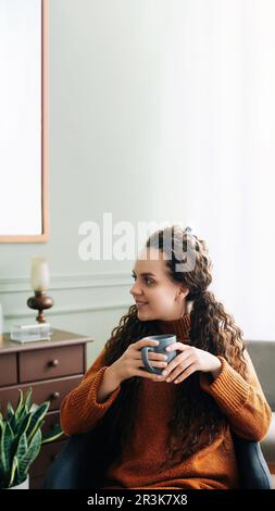 Traumhafte junge Frau, die warmen Kaffee in gemütlicher Inneneinrichtung genießt. Eine wunderschöne Dame, die ein heißes Getränk zu sich nimmt und sich zu Hause entspannt. Ruhige Frau mit warmem Becher Stockfoto