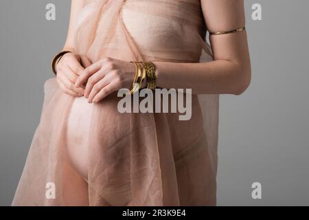 Kurzer Blick auf eine elegante und stilvolle schwangere Frau in weichem Chiffon-Stoff und goldenen Accessoires, isoliert auf grauem Hintergrund, Modekonzept für Umstandsmode Stockfoto