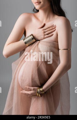 Zugeschnittene Ansicht einer stilvollen und eleganten schwangeren Frau, die in goldenen Accessoires und transparenten Chiffondrapierungen isoliert auf grauem Hintergrund posiert, Mutterschaft Stockfoto