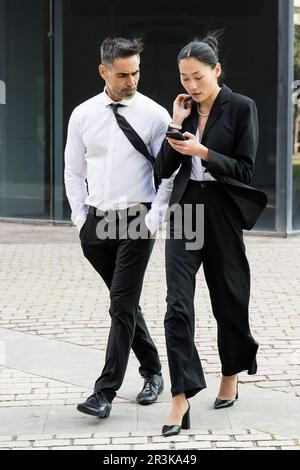 Geschäftsfrau und Geschäftsmann, die ein Telefon checken und auf der Straße laufen. Zwangloses Treffen zwischen einer jungen Führungskraft und einem Senior ceo während der SMS Stockfoto
