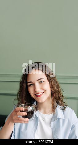Glückliche junge Hausfrau, die eine gemütliche Pause mit heißem Tee auf dem Sofa zu Hause genießt. Lächelnde Dame, die sich bei einem warmen Getränk entspannt und in die Kamera schaut Stockfoto