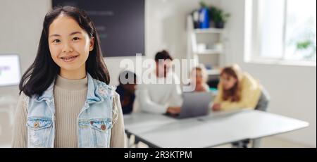 Ein selbstbewusster Schüler schaut in die Kamera, während der Lehrer andere Schüler im Hintergrund anspricht. Ein Mädchen, das an einer Schule digitale Kompetenz besucht. Stockfoto