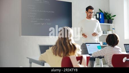 Männlicher Lehrer, der Kinder durch das Erlernen des Programmierprozesses führt. Vor der Klasse erklärt er die Grundlagen der Programmierung und Stockfoto