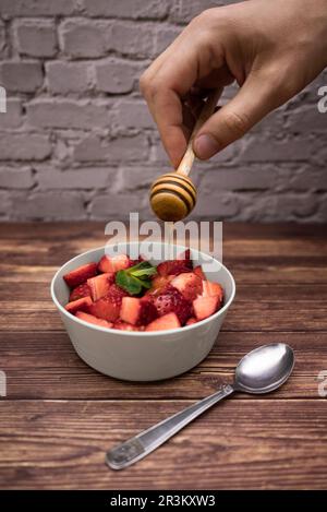Ein Mann, der Honig auf frische Erdbeeren schüttet. Kunststoffschüssel, Teelöffel, vertikales Bild, selektiver Fokus. Stockfoto