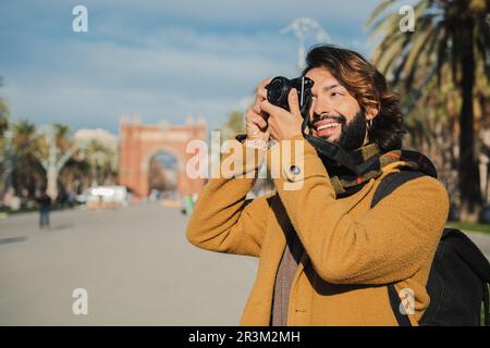 Glücklicher Touristenfotograf, der lächelt und ein Foto auf der Straße von Barcelona macht. Junger Reisender, der sich mit der Kamera auf eine Reise in Europa konzentriert. Ein bärtiger Mann genießt seine Freizeit am Wochenende. Hochwertiges Foto Stockfoto