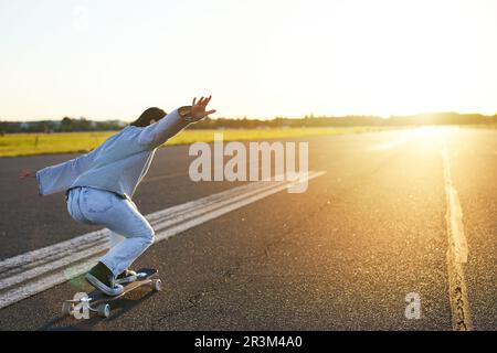 Glückliches Skateboard-Mädchen, das auf dem Skateboard fährt und Spaß auf der leeren Straße hat. Lächelnde Frau, die eine Fahrt auf einer sonnigen Straße genießt Stockfoto