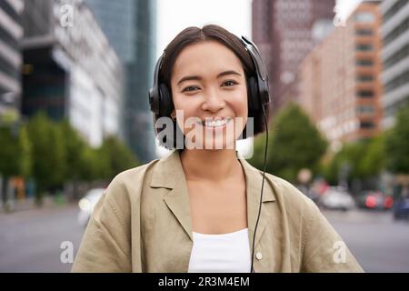 Porträt einer lächelnden asiatischen Frau in Kopfhörern, die im Stadtzentrum auf der Straße steht, glücklich aussieht und Musik hört Stockfoto