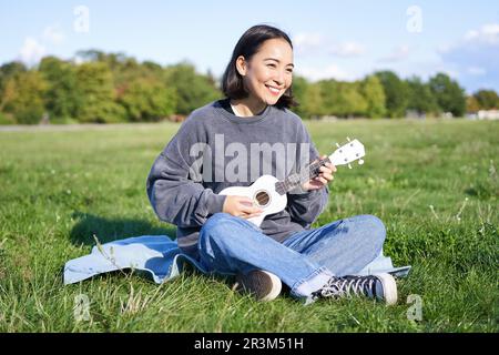 Wunderschönes asiatisches Mädchen, das im Park sitzt, Ukulele spielt und singt, sich an einem sonnigen Frühlingstag im Freien entspannt Stockfoto