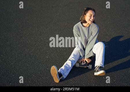 Glückliche, wunderschöne koreanische Teenagerin sitzt auf ihrem Skateboard, fährt auf Longboard und trägt lässige Kleidung Stockfoto