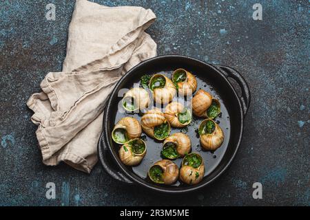 Escargots de Bourgogne kochte Schnecken mit Knoblauchbutter und Petersilie in schwarzer Gusseisenschale auf rustikalem Steinhintergrund von oben, traditionell französisch Stockfoto