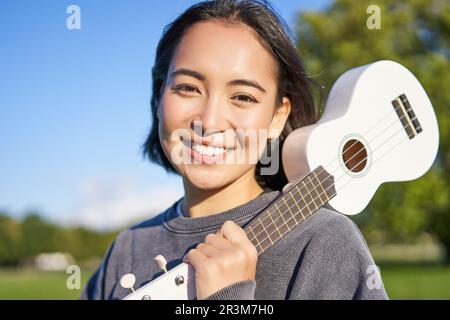 Portrait eines wunderschönen lächelnden Mädchens mit Ukulele, einer asiatischen Frau mit Musikinstrument, die sich draußen im Green Park posiert Stockfoto