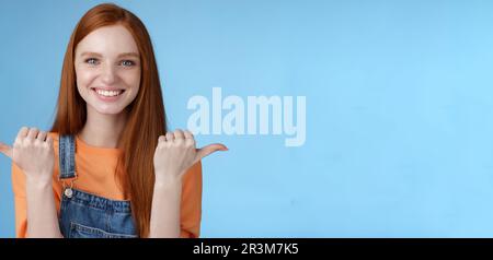 Innenaufnahme charismatisch durchsetzungsfähig glücklich lächelnd rothaarige Frau orange Hemd Denim Overalls zeigt seitwärts Daumen links rechts sh Stockfoto