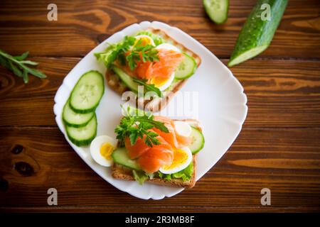 Gebratener Toast mit Salat, Ei, Gurken und rotem Fisch auf einem Teller, auf einem Holztisch. Stockfoto