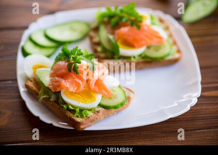 Gebratener Toast mit Salat, Ei, Gurken und rotem Fisch auf einem Teller, auf einem Holztisch. Stockfoto