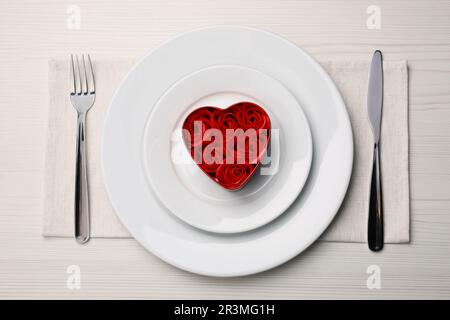 Schöne Tischgestaltung. Geschirr und dekorative herzförmige Box mit Rosen für romantisches Abendessen auf einem weißen Holztisch, flach liegend Stockfoto