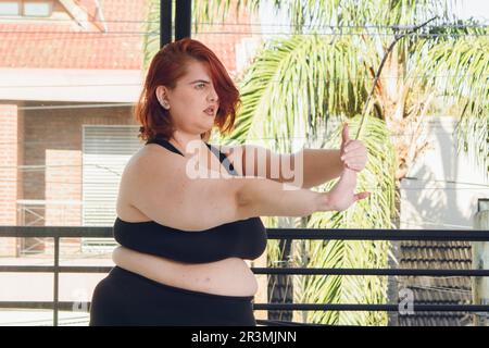 Junge, übergroße argentinische Latina-Frau mit kurzen roten Haaren, trainiert drinnen, streckt Arme und Hände, trägt schwarze Sportbekleidung. Stockfoto