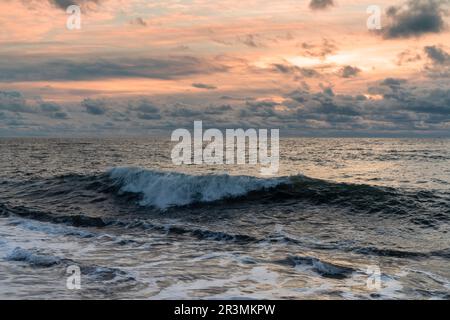 Hintergrundlandschaft einer großen Welle, die unter einem stürmischen Sonnenuntergang zum Ufer rollt Stockfoto