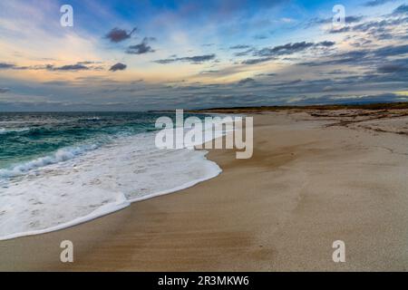 Ein niedriger Winkel Blick auf den weißen Sandstrand mit Wellen, die sich unter einem farbenfrohen Sonnenuntergang hineinrollen Stockfoto