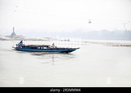 Traditionelles hölzernes Passagierboot auf dem Brahmaputra River in der Nähe von Guwahati mit einer Seilbahn darüber, die Guwahati mit anderen Flussufern in Assam, Indien verbindet Stockfoto