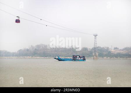 Traditionelles hölzernes Passagierboot auf dem Brahmaputra River in der Nähe von Guwahati mit einer Seilbahn darüber, die Guwahati mit anderen Flussufern in Assam, Indien verbindet Stockfoto