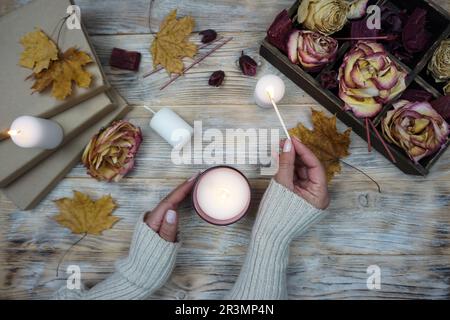 Eine Frau zündet eine aromatische Kerze an. Auf dem Tisch liegen getrocknete Rosen und gelbe Ahornblätter. . Hochwertiges Foto Stockfoto