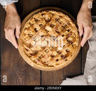 Zwei weibliche Hände halten einen runden gebackenen Kuchen mit Apfelfüllung auf einem braunen Holzbrett. Blick von oben Stockfoto