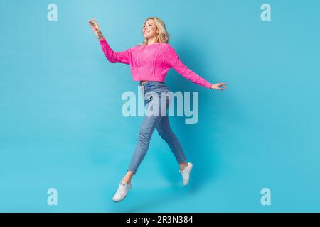 Der Kadre einer jungen, wunderschönen Frau trägt einen rosa Pullover mit Sprungschritten, unachtsamer Tragekomfort, isoliert auf blauem Hintergrund Stockfoto