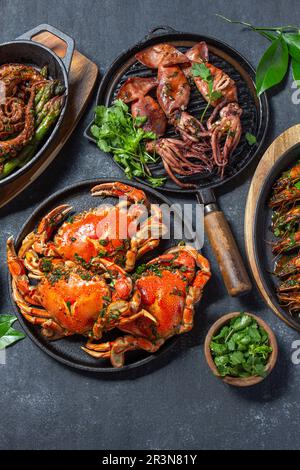 Auswahl an Meeresfrüchtegerichten. Krabben, Kraken, Tintenfische und Tigergarnelen auf gusseisernen Pfannen und Tellern auf schwarzem Hintergrund. Draufsicht. Stockfoto