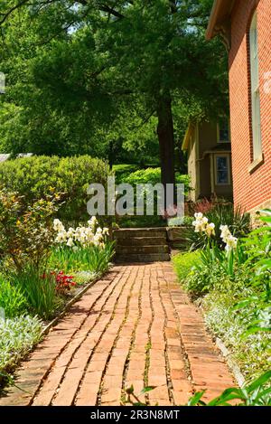 Ein alter Backsteinpfad führt durch einen hellen und farbenfrohen Garten neben einem alten Haus im historischen Dorf Roscoe. Stockfoto