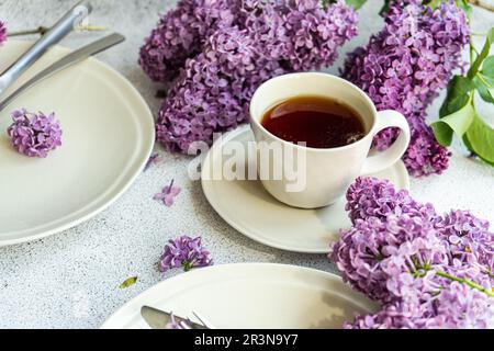 Vorderansicht mit weißer Tasse Tee, weißen Tellern, Messern und wunderschönen Fliederblumen auf grauem Tisch Stockfoto