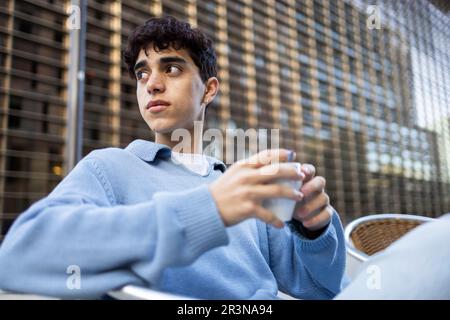 Nachdenklicher junger Mann mit dunklem Haar und blauem Pullover, der sich bei Tageslicht mit einer Kaffeetasse entspannen kann, während er auf ein verschwommenes Gebäude blickt Stockfoto
