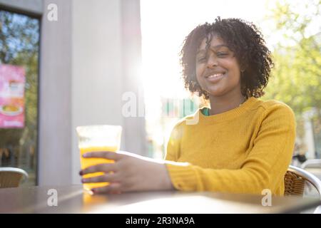 Positive junge afroamerikanische Frau mit lockigem Haar, die lächelt und in die Kamera schaut, während sie am Tisch im Café mit Orangensaft bei Tageslicht agai sitzt Stockfoto